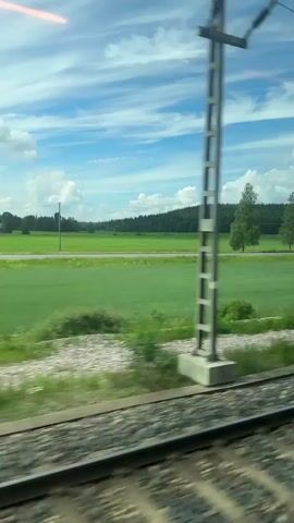 This is a video taken from inside a moving train, looking out the window. The landscape outside consists of green fields, trees, and a blue sky with white clouds. There are train tracks in the foreground, blurred due to the motion of the train. In the distance, there are also power lines and a few vehicles, including a white car and a motorcycle. The overall impression is of a scenic countryside view from a train journey.