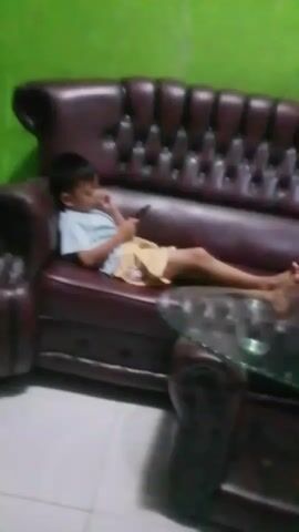 A young boy lies on a brown leather couch, looking at his phone. The couch is tufted, and the room has green walls and white tiled floors. A glass-topped coffee table is in front of the couch. The video is shaky and blurry, especially in the later frames.