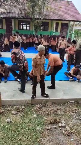 The video shows a group of school children sitting on blue tarps in an outdoor courtyard. They are dressed in uniforms. Some adults are present, including one man in a patterned shirt and dark pants who appears to be instructing or interacting with the children. Another man in a tan uniform is also visible. The background shows school buildings and trees. The ground is a mix of concrete and dirt/grass. Some children are looking at the adults, while others are looking down. The overall scene appears to be some kind of school activity or gathering.