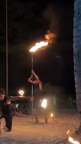 The video shows a nighttime fire show on a beach. Two men are the main performers, each holding flaming torches. One performer holds a tall torch that shoots flames high into the air. The other holds two smaller torches, one in each hand, manipulating them in the air. Both performers are kneeling on the sand. There are other people standing around watching the show. Palm trees are visible, some decorated with lights and cloth. The environment is dark with the torches illuminating the beach area.