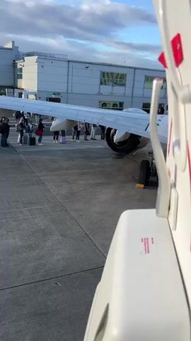 The video shows an airplane wing and engine on the tarmac of an airport. Several passengers are visible standing in a line next to the plane, holding luggage, and appearing to be boarding. The airport terminal building is in the background. The lighting suggests it is either early morning or late afternoon.