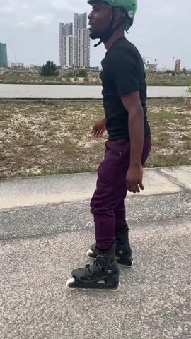 A Black man wearing a black t-shirt, purple pants, a green helmet, and black rollerblades is rollerblading on a paved path. He is in a park-like setting with dry grass and some trees. In the background, there are several tall buildings. The sky is overcast. The man begins by looking forward and then bends his knees and extends his arms. He then leans forward and bends his knees further, appearing to be preparing for or performing a trick. He then returns to a more upright position.