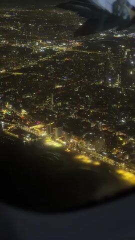 This is an aerial view from an airplane window at night. The scene shows a sprawling city illuminated by countless lights. The lights create a dense, textured pattern across the landscape, suggesting a heavily populated urban area. Along the coast, a line of brighter lights is visible, possibly indicating a waterfront promenade or a strip of businesses. A portion of the airplane's wing is visible in the upper left corner of the frame, framing the cityscape. The overall impression is one of a vibrant, active city at night.