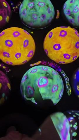 The video displays a close-up, vertical shot of a collection of colorful, orb-like objects against a dark background. These objects are decorated with intricate patterns of bright colors, including yellow, orange, pink, purple, and green. They appear to be projected images or illuminated surfaces, as a hand and fingers are seen interacting with them. The hand, covered in what appears to be a glove or sleeve, touches and touches the surfaces of these orbs, causing some of the projected light patterns to distort or change. The overall impression is one of a digital art installation or an interactive display that reacts to touch.