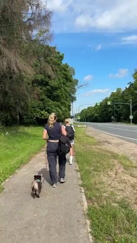 Two women and a dog are walking along a paved path next to a road. The woman in front is wearing a dark blue crop top and dark gray sweatpants. She is holding a leash attached to a small, fluffy gray and black dog. The woman behind her is wearing a white t-shirt and dark pants, carrying a black backpack. They are walking away from the camera, towards a road lined with trees. The sky is blue with scattered white clouds. The path is bordered by green grass and trees on the left and the road on the right. There are traffic lights visible in the distance.