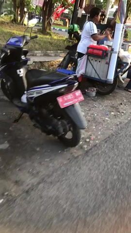 The video shows a street scene. In the foreground, a dark blue and white motorcycle is parked on a paved surface. The motorcycle has a red license plate that reads "S 1085 WAR". Behind the motorcycle, a person in a white t-shirt is standing at a food cart. The food cart has a silver container and a red insulated bag. The person at the cart appears to be interacting with someone off-camera. The background includes trees, a sidewalk, and a street with other vehicles. The lighting suggests it is daytime, possibly late afternoon or early evening. The scene is somewhat blurry, indicating movement during recording.