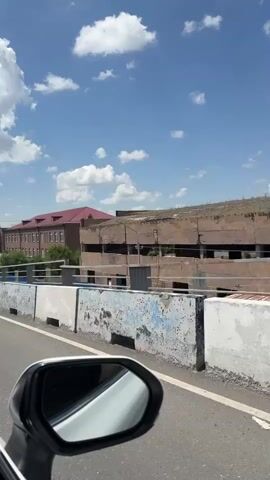 The video is shot from the passenger seat of a moving vehicle, looking out the side window. The perspective is slightly angled, with the side mirror of the car visible in the foreground on the left. Through the window, a bridge railing with grey posts and a blue band runs horizontally across the middle ground. Beyond the bridge, several buildings are visible. The closest building on the right appears to be under construction or derelict, with exposed concrete and rebar. To the left of that, a larger, older building with a red-tiled roof and multiple windows stands prominently. Further to the left, another multi-story apartment building with a beige facade and numerous windows is visible. Green trees are scattered between the buildings and behind the bridge railing. The sky above is bright blue with fluffy white clouds. The road in front of the bridge appears to be asphalt.