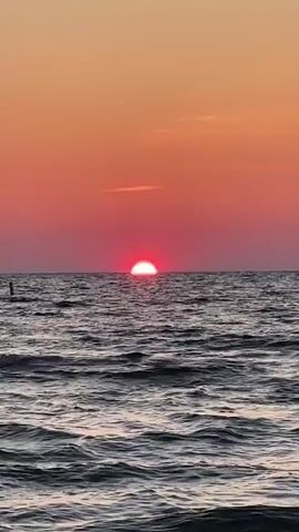 The video shows a sunset over the ocean. The sun is a bright red orb on the horizon, half submerged in the water. The sky is a gradient of orange and pink. The ocean is choppy with small waves. There are no people or man-made objects visible in the scene.