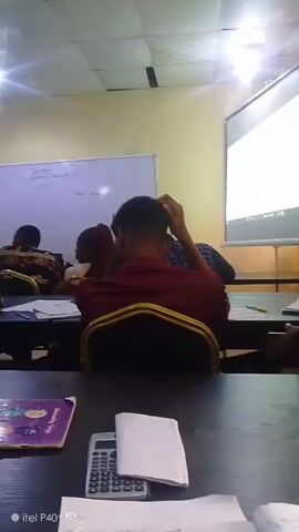 The video shows a classroom setting with students seated at desks. A projector screen is visible at the front of the room, displaying content. Several students are in view, some with their backs to the camera, others in profile. One student in the foreground is wearing a pink shirt and appears to be writing or interacting with something on the desk. A calculator and notebooks are also present on the desks. The lighting suggests an indoor environment, possibly a lecture or study session.