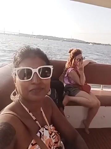 Here is a description of the video:
The video, shot on a boat, features a woman in sunglasses and a bikini top holding the camera and facing it. In the background, a man stands near the edge of the boat looking out at the water. Another woman, wearing a white hat and a black tank top, is taking a photo of the man. The boat has brown benches and a wooden ceiling with recessed lights. The scene is set on a sunny day with a view of the sea and distant land. Music plays in the background.