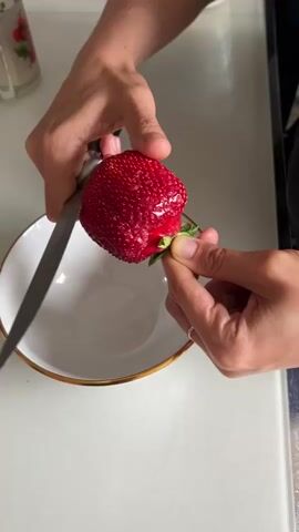 A person is cutting a strawberry over a white bowl with a gold rim. The person's hands are visible, holding the strawberry and a knife. The strawberry is being cut into slices, which are falling into the bowl. The background is blurry, but appears to be a kitchen or dining area.