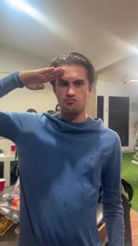 Here's a description of the video:

A young man in a blue hooded sweater is the focus of the video. Initially, he's looking directly at the camera with a serious expression, his hand raised in a salute. The background reveals a party atmosphere, with other people milling around. There are red cups and snacks on a table nearby, suggesting a casual gathering. A doorway leads to an outdoor area with artificial grass. As the video progresses, the man's serious expression melts into a smile.