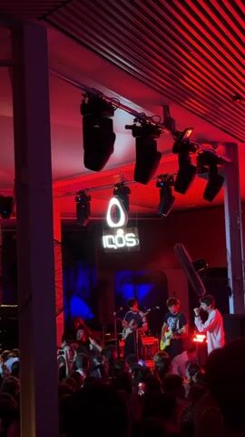 A band performs on a stage illuminated by red and blue lights. A crowd of people watches the performance. There are speakers and lights mounted above the stage. A neon sign that reads "IQOS" is visible behind the band. The video is shot from the perspective of someone in the audience.