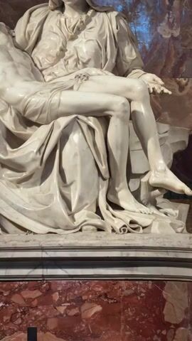 A medium shot features Michelangelo's Pietà sculpture, depicting Mary holding Jesus, made of white marble. Mary, clad in flowing robes, sits with a sorrowful expression, cradling Jesus' body across her lap. Jesus' head is tilted back, his left arm draped across Mary's leg, and his right arm hangs beside his body. His body shows visible wounds on his right side and feet. The sculpture rests on a multi-tiered base, the lowest layer being a reddish-brown marble with white veins, and the top layer a plain white marble. A reddish-brown, patterned wall is visible behind the sculpture, and a small, blue-tinted window is visible in the top background.