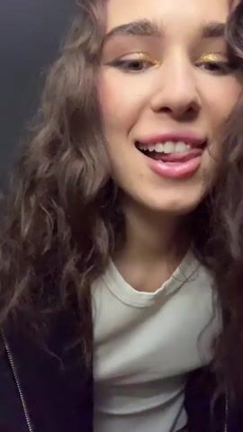 A young woman with curly brown hair and gold eyeshadow is dancing. She is wearing a white t-shirt under a black jacket. She is looking at the camera and sticking her tongue out. She gives two thumbs up, then makes a “blow kiss” gesture towards the camera. She does a mock-serious, scowling face, then turns around and strokes her hair. The background is a dark grey or black wall. Upbeat pop music is playing.