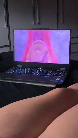 A laptop is open on a bed, displaying an anime scene. The laptop has a black keyboard with illuminated blue keys. In the scene, a character with pink hair and green clothing is visible. The room appears to be a bedroom with a wardrobe in the background. The lower part of the frame shows a person's legs.