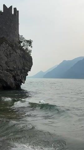 The video shows a serene lake with a large, rocky cliff on the left side, topped with a section of an ancient stone structure resembling a castle turret. The water is choppy, with waves breaking near the shore. In the middle ground of the lake, a white swan is gracefully swimming. Further in the distance, a dark-colored duck can be seen. The background is dominated by the hazy outlines of large, majestic mountains stretching across the horizon under a pale, overcast sky. The overall scene evokes a sense of natural beauty and historical presence.