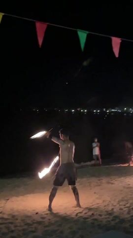 At night on a beach, a man is performing a fire dance. He is shirtless and wearing a loincloth, holding two fire batons. He is performing in front of a crowd of people who are watching. There are colorful triangular flags strung up above, and the city lights twinkle in the background. Another fire dancer is visible to the right, also holding batons. The beach is sandy, and the waves are gently lapping at the shore.