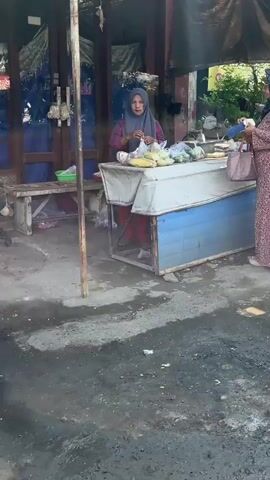 The video shows a market scene. A woman wearing a hijab and a pink top is standing behind a stall filled with vegetables, including corn. She is holding a plastic bag and appears to be packaging some produce. Another woman, wearing a patterned dress, stands to her right, holding a shopping bag. The stall has a blue and white counter. The ground is uneven and appears to be wet in some areas. In the background, there are stalls with tarps and hanging items. The overall environment suggests an outdoor market setting.