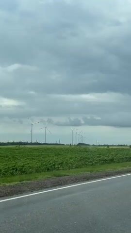 This media file captures a road trip scene in Zarevo, Russia, under a dramatic and cloudy sky. The primary subjects are a series of wind turbines scattered across a vast agricultural landscape. These tall, white structures with their characteristic blades are positioned in the distance, suggesting a wind farm.

The scene is set alongside a paved road, indicated by the asphalt surface and white lane markings visible in the foreground. To the side of the road, green fields stretch out, likely crops, with some areas showing ploughed earth. The weather appears to be overcast, with heavy, grey clouds dominating the sky, though there are glimpses of blue sky in some frames, and patches of sunlight break through. This dynamic weather suggests a possibly unsettled or changeable day.

The activity taking place is travel, as indicated by the perspective from a moving vehicle. The camera is positioned to capture the passing scenery, giving the impression of a journey through the countryside. There are no people or animals visible in the frames. The overall mood is somewhat somber due to the cloudy weather, but the presence of the wind turbines offers a hint of modern infrastructure within the rural setting. The location cues, such as the wind turbines and the specific mention of Zarevo, Russia, help to contextualize the imagery. There is no visible text within the media file.