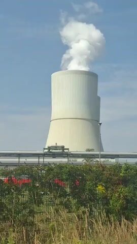 The video shows a panoramic view of an industrial facility on a clear day with a blue sky. The most prominent structure is a large, white, hyperboloid cooling tower emitting a plume of white steam. To the left of the cooling tower are two gray, rectangular buildings, also emitting plumes of steam from their upper sections. In the foreground, there is a chain-link fence with tall dry grass and some green bushes and plants with red and yellow flowers. Behind the fence, there is a section of pipes supported by elevated structures. Further in the background, within the facility, various industrial equipment and structures are visible. The camera pans from right to left, showcasing different parts of the plant.