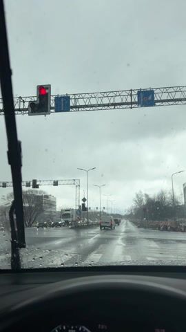 The video shows a view from inside a car stopped at a red light on a rainy day. The windshield wipers are in motion, clearing raindrops. The road is wet and reflects the surrounding lights. Traffic signals and street signs are visible overhead. Other vehicles are present at the intersection. The car's dashboard is visible, showing a temperature reading of 7.5 degrees Celsius and a time of 15:13. Construction or road work appears to be taking place on the right side of the road.