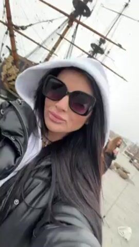 A woman with long black hair and large sunglasses is taking a selfie in front of a large wooden ship with tall masts. She is wearing a black leather jacket and a white hoodie. She adjusts the hood with her right hand. The sky is overcast. Another woman is visible walking past in the background.