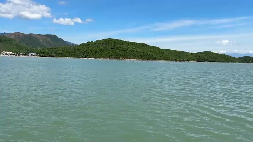 The video is a wide shot of a calm turquoise sea under a bright blue sky with scattered white clouds. Lush green hills and mountains form the coastline in the background, dotted with a few small buildings. The camera appears to be moving slightly, creating a gentle ripple effect on the water's surface. The overall impression is one of a serene coastal landscape.