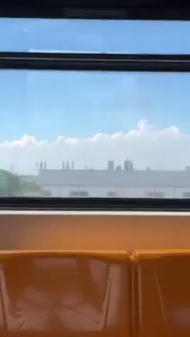 The video shows a view from inside a train. The camera looks out of the train window at the city skyline in the distance. The skyline is faint, and clouds fill most of the sky. The train is moving, as the view outside changes. There is a large white water tower that comes into view. The seats inside the train are orange.