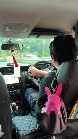 The video captures the interior of a car from the backseat perspective, focusing on the driver. The driver, who appears to be a young person with dark hair, is seen from behind, gripping the steering wheel. They are wearing a dark shirt and blue jeans. A pink plush bunny keychain is attached to a black bag hanging from the back of the passenger seat, obscuring part of the driver's leg. In the foreground, a pamphlet with a 7-Eleven logo is visible in a seat pocket. The car's dashboard displays a screen, and a smartphone is mounted on a holder on the dashboard. Outside the car, trees and road signs are visible, indicating the vehicle is on a road with some curves. The lighting suggests it is daytime.