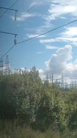 A video shows a view of a power substation with tall lattice towers and overhead power lines against a bright blue sky with scattered clouds. Vegetation, including trees and bushes, is visible in the foreground and surrounding the substation. The camera appears to be moving, possibly from a vehicle, as indicated by the slight blur and shifting perspective. There are no people visible in the footage.