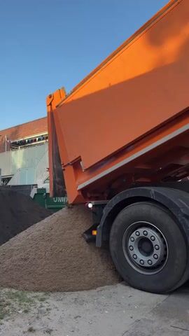 A side view of an orange dump truck is shown as it unloads a large pile of light brown sand onto the ground. The sand is spilling from the rear of the truck's bed, creating a large mound. In the background, a dark pile of material, possibly soil or another type of dirt, is visible, along with parts of a building and a clear blue sky. The truck's large rear wheel and part of its chassis are also in the frame. The lighting suggests it is either early morning or late afternoon. There are no people visible in the video.