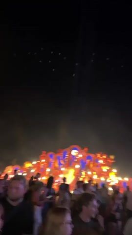 The video shows a crowded outdoor event at night. In the foreground, a crowd of people are visible, some looking towards the camera, others interacting. In the background, a large, elaborately decorated stage or structure is illuminated with bright lights, primarily orange and blue. There is smoke or fog in the air, catching the light and creating an atmospheric effect. In the sky above, small green lights are scattered, and a bright light beam is visible originating from the stage area and pointing upwards. The overall impression is of a lively festival or concert.