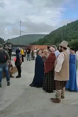 The video captures a group of people dressed in historical attire, possibly a reenactment or a film set. In the foreground, a man with a beard wears a light-colored shirt, a beige vest, brown pants, and a cap. He stands next to a woman in a blue dress and a white headscarf, and another woman in a maroon shawl over a plaid dress and a head covering. A woman in a dark blue cape is also visible. Behind them, other individuals and some large shipping containers are present. The background shows a forested hillside under an overcast sky. The overall impression is that of a staged historical scene in an outdoor location.