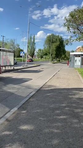 The video shows a street scene with a sidewalk and a road. On the sidewalk, there is a bus stop with a shelter and advertisements. A white van is parked at the bus stop. Across the street, there is a pedestrian crossing with a traffic signal. Several trees and streetlights are visible along the road. A person in a red shirt is walking across the street. The sky is blue with some clouds.