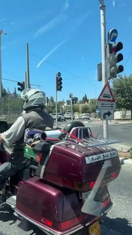 A man wearing a white helmet and a grey vest is riding a red motorcycle. He has a long white beard and is wearing a light-colored shirt and dark pants. The motorcycle is loaded with items, including a green box with tomatoes and a patterned rug. The motorcycle is stopped at a traffic light, which is red. There are traffic signs in the background. The environment appears to be an urban street with buildings and trees. The sky is blue.