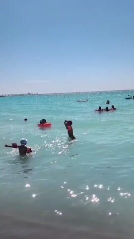A sunny day at the beach shows several people enjoying the clear, turquoise water. In the foreground, a child wearing a white hat and arm floaties is facing left, splashing in the shallow water. To the right of this child, another child, also wearing arm floaties, stands facing forward and moves their arms. Further out in the water, a person in an orange float is visible. In the middle distance, a group of at least three people are together in the water, some appearing to be in red floatation devices. In the background, more people are spread out in the water, and a distant bridge or structure is visible on the horizon under a clear blue sky. The water appears calm with some gentle ripples and sunlight reflecting off the surface. The shore is dark and out of focus in the very bottom of the frame.