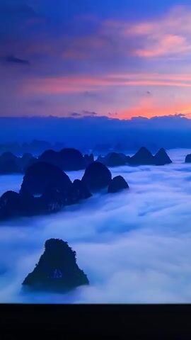 The video shows a serene, aerial view of karst peaks emerging from a sea of clouds at sunrise. The sky displays a vibrant palette of blues, purples, and oranges as the sun ascends. The mountains are silhouetted against the colorful sky, with a thick layer of white clouds filling the valleys between them, creating a dreamlike landscape. The camera slowly pans across the scene, offering a panoramic perspective of this natural wonder.