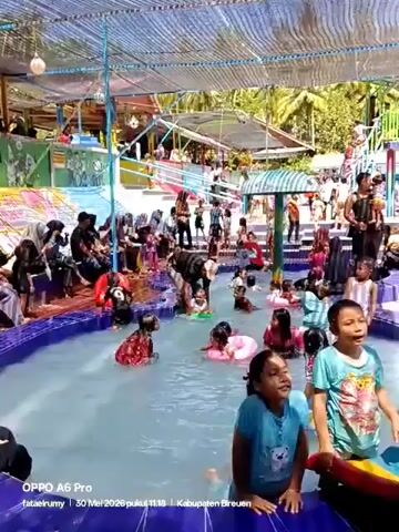 The video shows a crowded swimming pool with many people, mostly children, enjoying themselves. Some children are in the water, some with inflatable rings, while others are sitting or standing on the edge. Several adults are also present, some watching over the children. The pool is circular with a blue-tiled edge. The pool area is covered by a metal structure that provides shade. Banners and decorative elements are present. The video's text indicates it was shot with an OPPO A6 Pro in Kabupaten Bireuen.