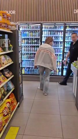 A family, a man, a woman, and a young girl, walk past refrigerated drink and wine displays in a store. The woman walks in front, her back to the camera, wearing a light-colored jacket and jeans. The man follows her, wearing a black shirt and dark pants. The girl, dressed in a pink fuzzy jacket and a sparkly dress, walks beside the man, holding a white stuffed animal. The store has rows of bottled drinks and wine bottles on display.