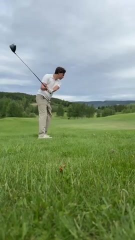 A young man in a white shirt and tan pants is seen golfing on a green golf course. He swings his golf club and watches the ball. He then walks after it, holding his golf club. The course is surrounded by trees and rolling hills under a cloudy sky.
