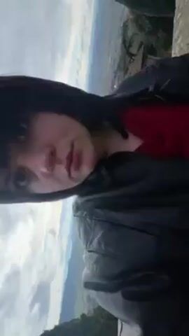 The video shows a person, presumably a young woman, in what seems to be an elevated location, possibly a viewpoint in a rural setting. She has short dark hair with bangs, a fair complexion, and is wearing a black leather jacket over a red top. She is looking into the camera, and the perspective suggests she is holding the camera herself, potentially taking a selfie video. 

Behind her is a panoramic view of the landscape. The landscape includes a small settlement with red-tiled roofs, tall, slender trees, and a vast expanse of green fields stretching into the distance under an overcast sky. The camera angle is slightly tilted to the left, giving a broad view of the surrounding area. The scene is set outdoors on a cloudy day.