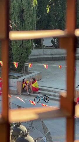 The video captures a candid moment in Las Rozas de Madrid, Spain, viewed through a wooden lattice. Two young individuals, draped in Spanish flags, are seated on stone steps in what appears to be a park or public square. Their body language suggests a moment of quiet conversation or shared contemplation. One person is leaning against the steps, while the other is seated next to them, facing inwards. A small bicycle with training wheels is parked nearby, suggesting the presence of children enjoying the outdoors.

The scene is bathed in warm, natural light, indicating it's likely late afternoon or early evening. The surrounding environment features lush green trees and more Spanish flags strung across the area, hinting at a festive atmosphere or a public event related to Spain. The overall mood seems peaceful and somewhat introspective, despite the celebratory context implied by the flags. The image is framed by the wooden bars of a window or gate, adding a sense of depth and perspective to the scene. No visible text is present within the frame.
