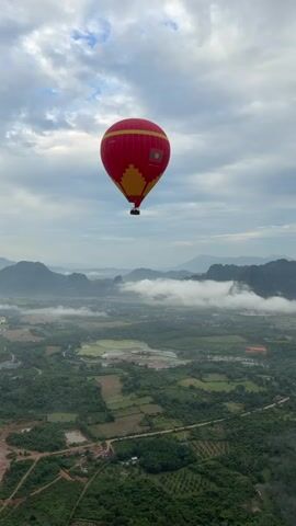A red hot air balloon with a yellow geometric pattern on its side and a flag logo on the top is suspended in the sky against a cloudy sky. Below the balloon, a vast landscape unfolds, featuring rolling green hills, lush forests, and patchwork fields. Some areas appear to be farmland with visible irrigation systems. In the distance, misty mountains rise, partially obscured by clouds. A scar-like feature, possibly a quarry or landslide, is visible on the side of a mountain to the left, showing exposed rock and reddish-brown soil. The overall scene depicts a serene aerial view of a rural landscape under a dynamic sky.
