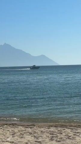 The video shows a beach scene with a clear blue sky and calm ocean waves. In the distance, a boat is sailing on the water, leaving a wake behind it. Mountains are visible in the background, partially obscured by haze. The foreground features a sandy beach. The video is very short, only four seconds long, and the boat is the only moving object. There is one person visible in the water.