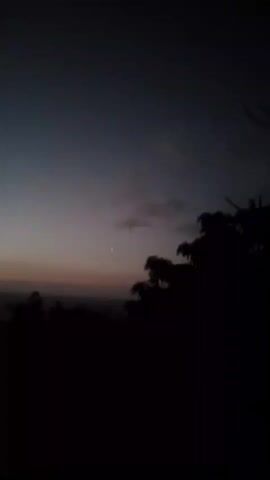 A video of a dusky sky with a single bright star and silhouetted trees. The sky is a gradient of dark blue to a faint orange near the horizon. There are a few wispy clouds. The trees in the foreground are dark and obscure most of the landscape below. The video appears to be taken at dusk or dawn.