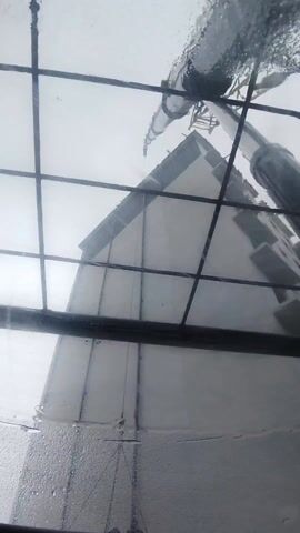 From the perspective of inside the cab of a crane on a rainy day, a platform carrying several workers is being lifted alongside the exterior wall of a tall, white building. The platform is suspended by a chain and cable rigging system. The interior of the cab contains various screens and controls visible, including what appears to be a GPS or guidance system on a tablet. The windshield is wet and the wiper is briefly visible in the first frame. The video ends showing the workers being lifted along the side of the building.