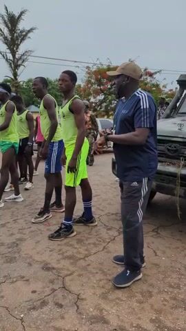 A group of athletes in running attire are lined up, appearing to be preparing for a race or training session. Most are wearing light green tank tops and shorts of various colors. A coach or official is standing to the side, wearing a navy blue Adidas jacket and grey track pants, holding a device that could be a timer. The surface they're on appears to be cracked concrete or asphalt. There's a sign and vehicles in the background. The weather seems overcast. One athlete is stepping forward, seemingly ready to begin.