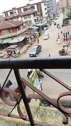 The video shows a street scene in what appears to be a developing country, viewed from an elevated perspective behind a railing. A gray pickup truck is positioned on the left side of the street, facing the viewer. A silver car is in front of it, moving away. On the left side of the street are informal structures resembling stalls or shops with a variety of goods on display. A woman in a pink striped shirt is walking away from the shops with a child in a pink dress. Other pedestrians are visible along the street. The street appears to be paved but possibly in disrepair with visible drains. There are several buildings of varying heights on both sides of the street. A sign for "Reality Max" is visible in the foreground. A yellow tricycle taxi is visible on the right side of the screen in the foreground. The sky appears overcast.