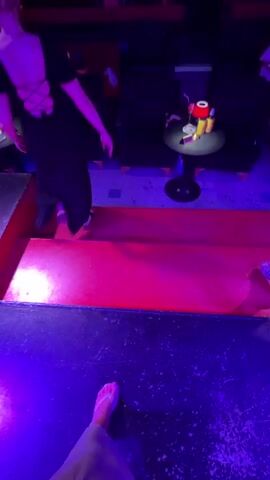 A person films themselves with a smartphone while standing on a stage. The stage is lit with purple and blue lights and has a reflective, glitter-like backdrop. There are red curtains on either side of the stage, and a disco ball hangs from the ceiling. Two other people are visible in the background, one on the left wearing a black dress and another on the right wearing a blue shirt and khaki shorts. The person filming is wearing a white bikini top and a flowing white skirt. They smile at the camera and raise their left arm.